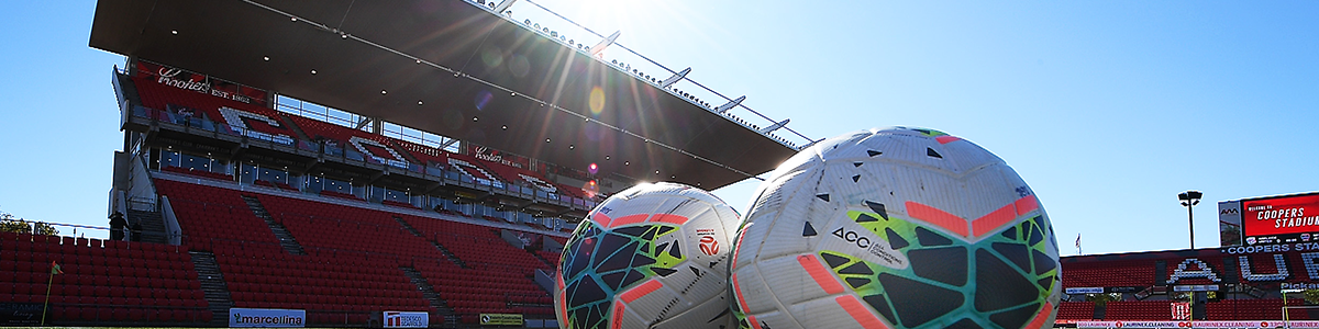 Two footballs on the pitch with a Hindmarsh Stadium stand in the background